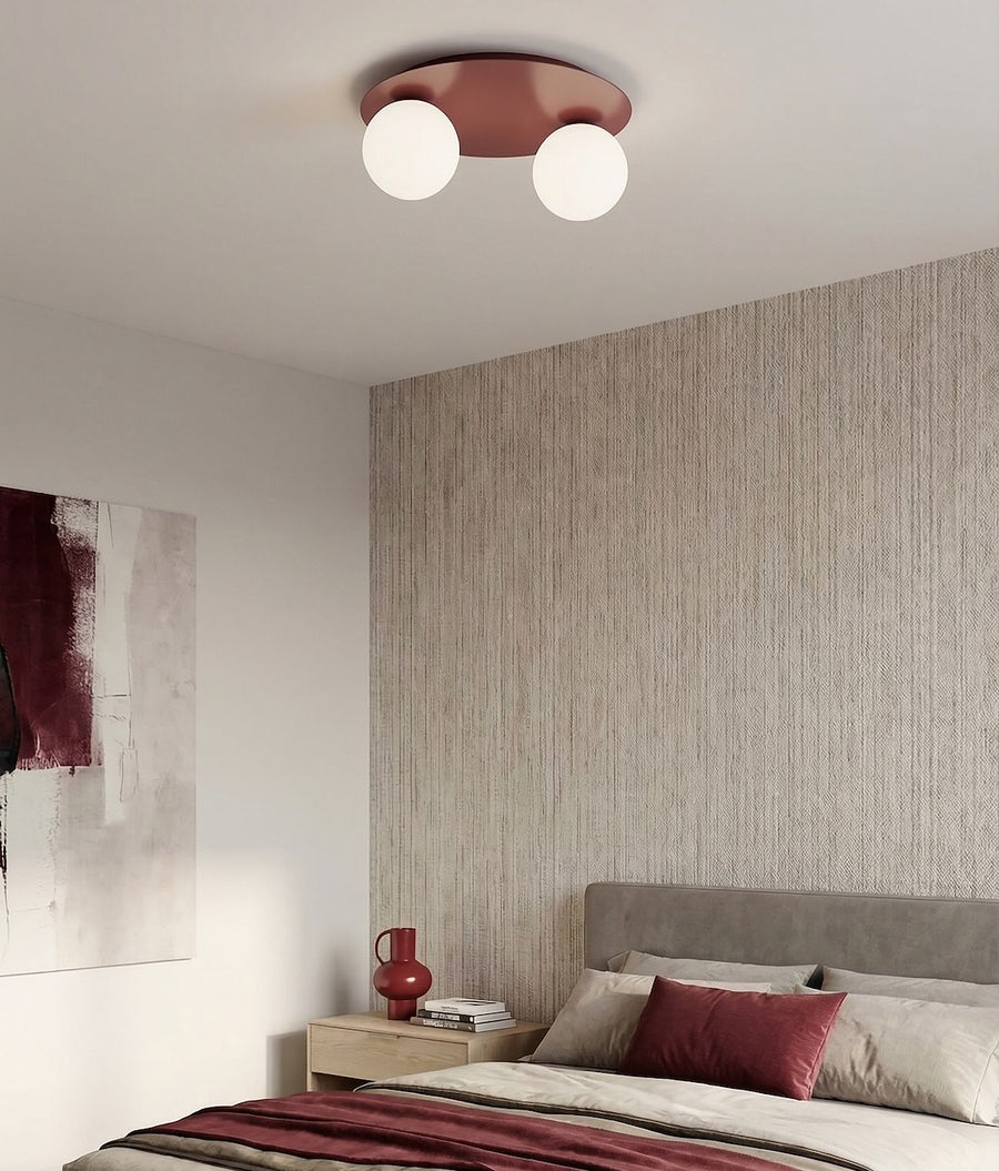 Ambiance de chambre avec papier peint texturé, plafonnier bordeaux, lit gris et coussins rouges.