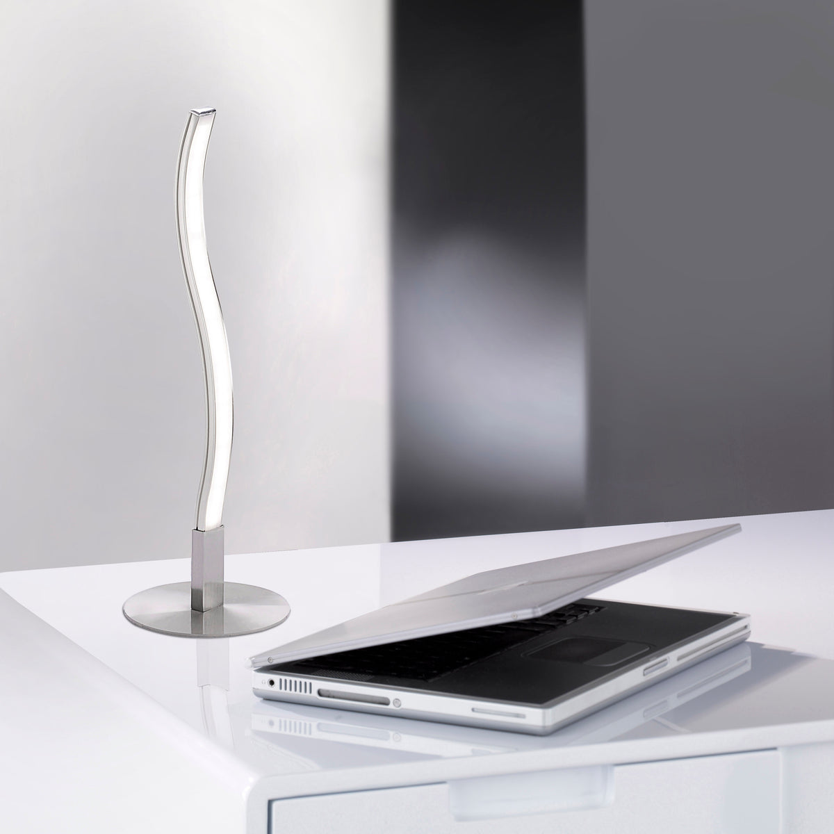Lampe à poser argentée posée sur un bureau blanc, près d'un ordinateur portable ouvert.