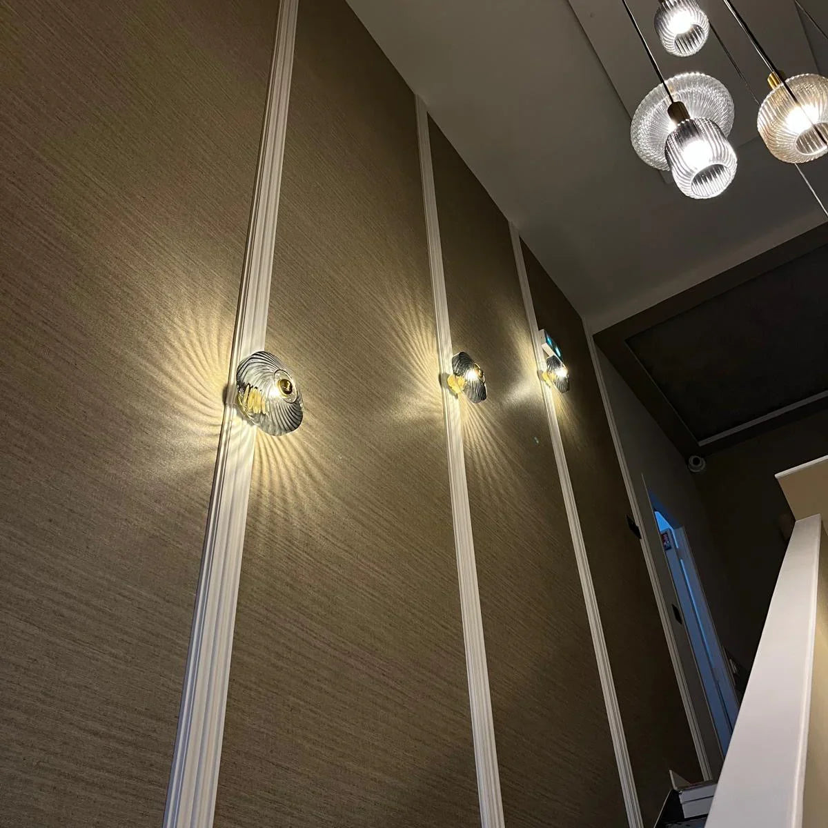 Trois appliques transparentes alignées sur mur marron, suspensions visibles en arrière-plan