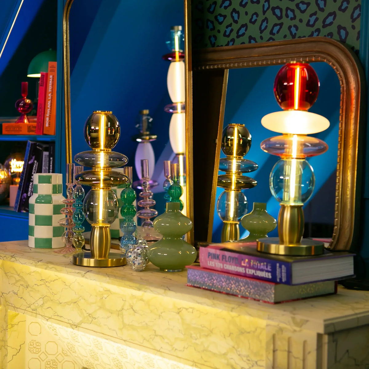 Lampe multicolore posée sur des livres, devant un miroir et un mur bleu, décor coloré.