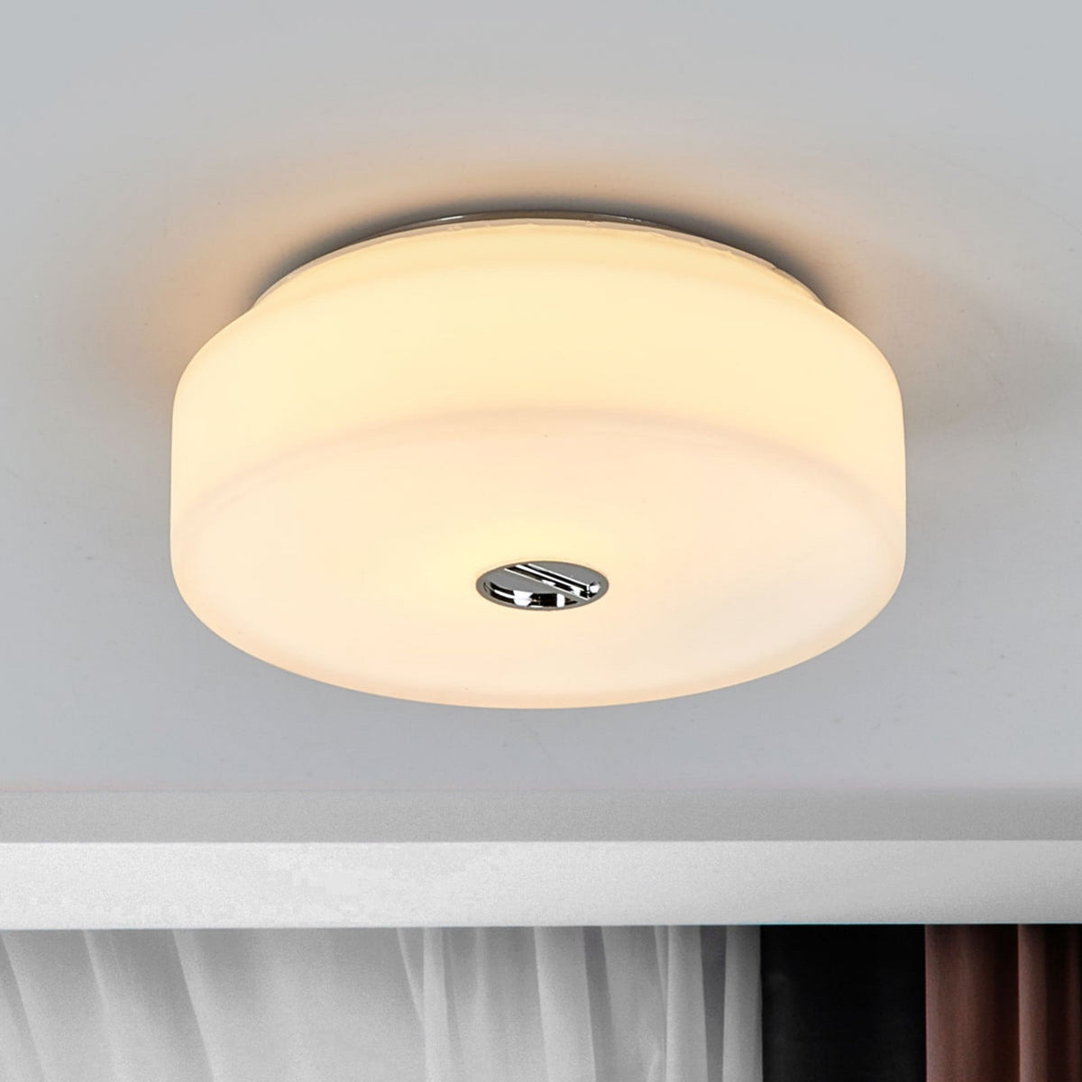 Plafonnier rond allumé, fixé au plafond, diffuseur en verre blanc et bouton central chromé.