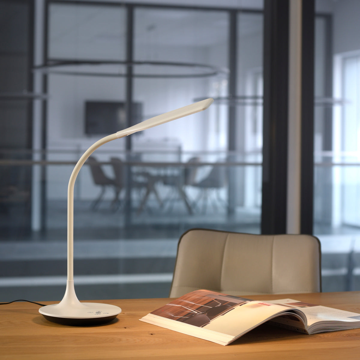 Lampe posée sur table en bois, magazine ouvert, bureau vitré en arrière-plan flou.
