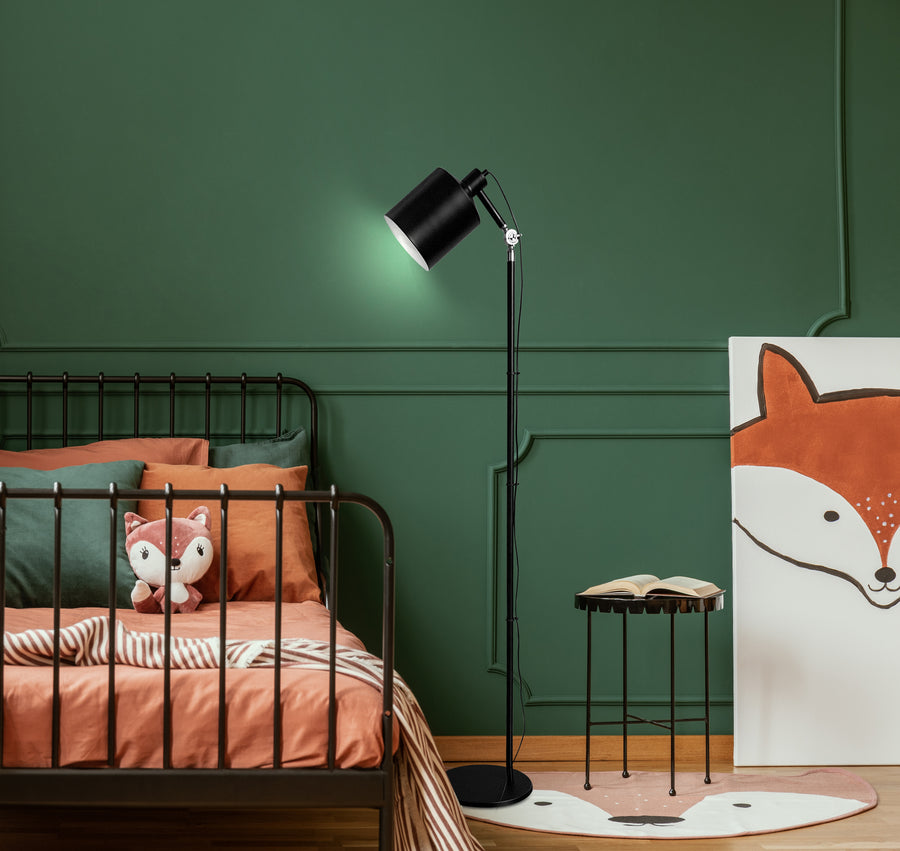Lampadaire noir allumé dans une chambre verte, près d'un lit et d'une petite table.
