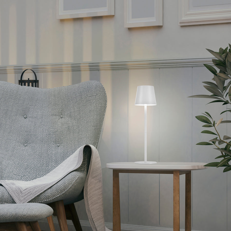 Lampe blanche allumée posée sur une table d'appoint près d'un fauteuil gris.