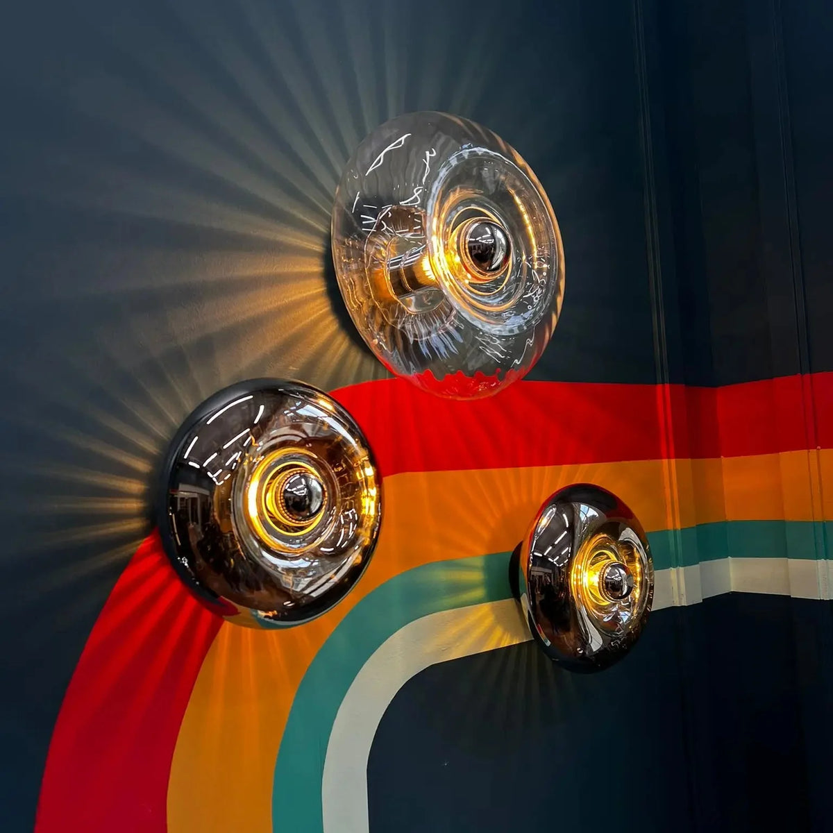 Trois appliques en verre fumé et transparent devant une peinture murale multicolore