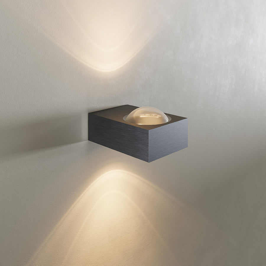 Vue de face sur mur blanc, applique noire brossée diffusant un éclairage haut et bas.