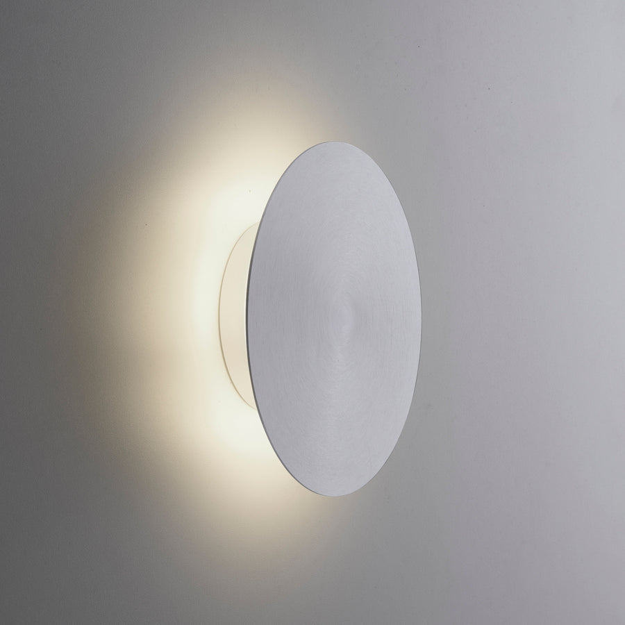 Vue rapprochée de l'applique aluminium allumée sur un mur, diffusant une lumière chaude avec halo.