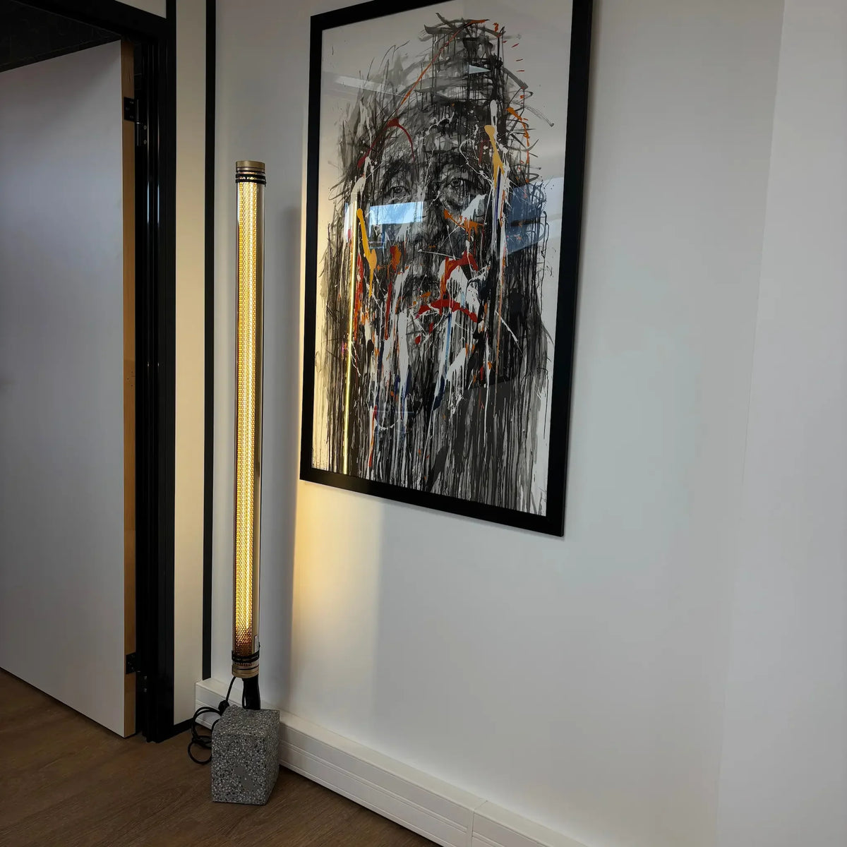 Lampadaire doré allumé contre un mur blanc à côté d'une peinture abstraite.