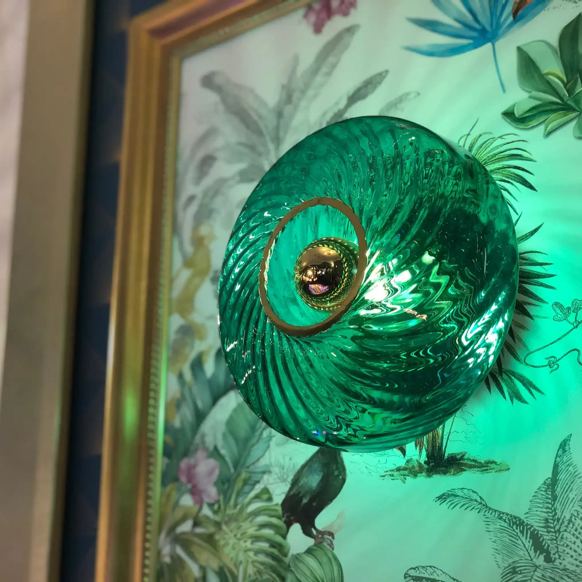 Gros plan applique en verre émeraude brillant devant un papier peint floral tropical