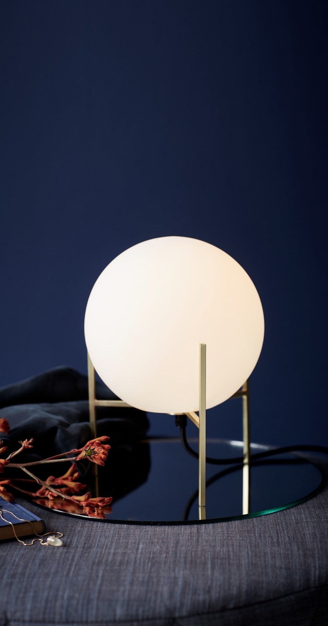 Lampe allumée posée sur une table en verre, tissu sombre et fleur séchée.
