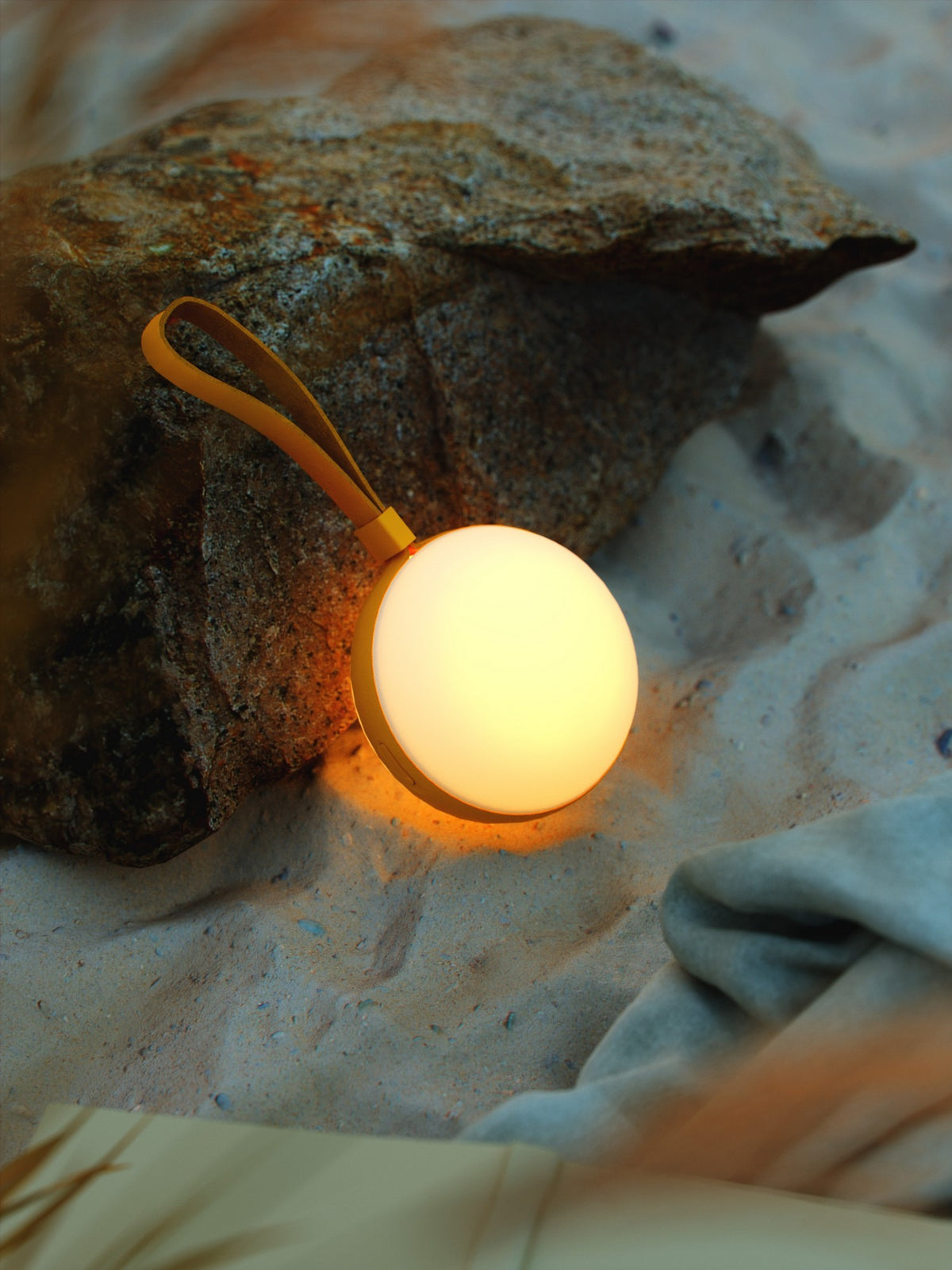 Mise en ambiance : lampe jaune allumée sur le sable, rocher et tissu.