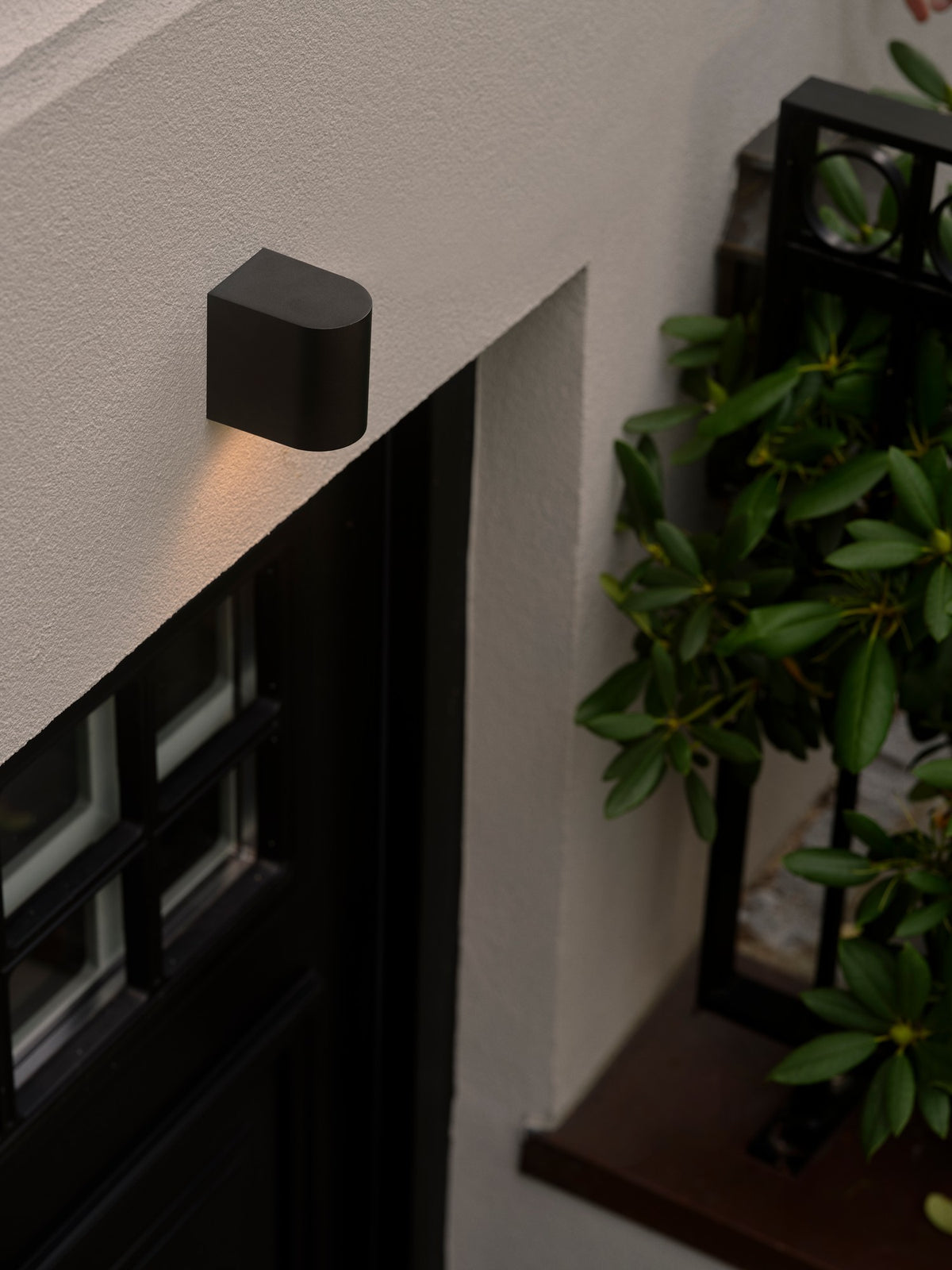 Mise en ambiance extérieure : applique noire allumée au-dessus d'une porte-fenêtre, plantes.