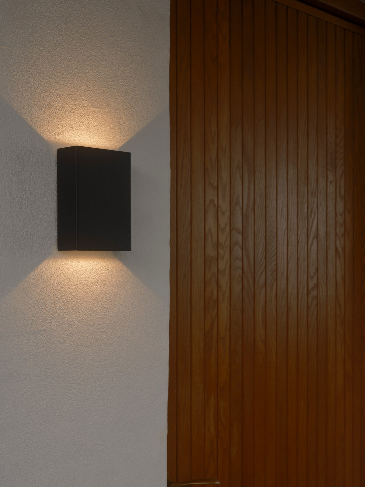 Gros plan d'une applique murale noire allumée sur mur blanc, près d'un bois clair.