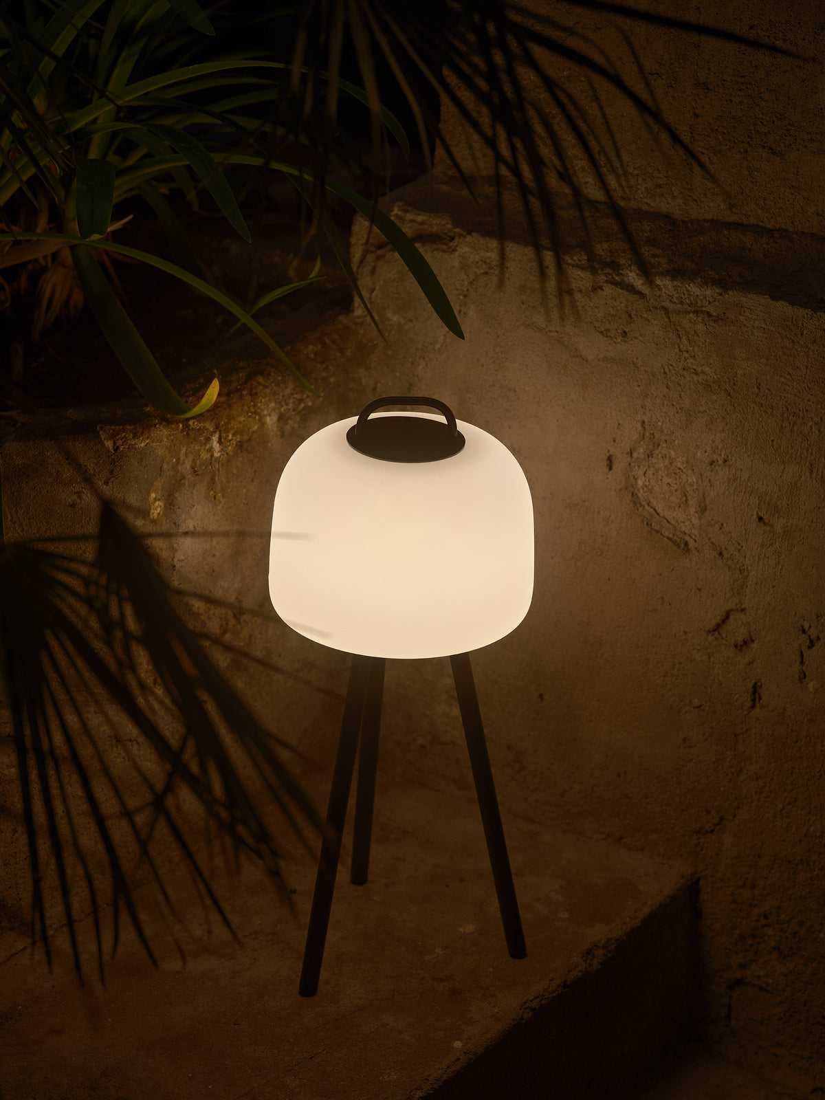 Lampe allumée fixée sur un trépied noir, près d'un mur extérieur.