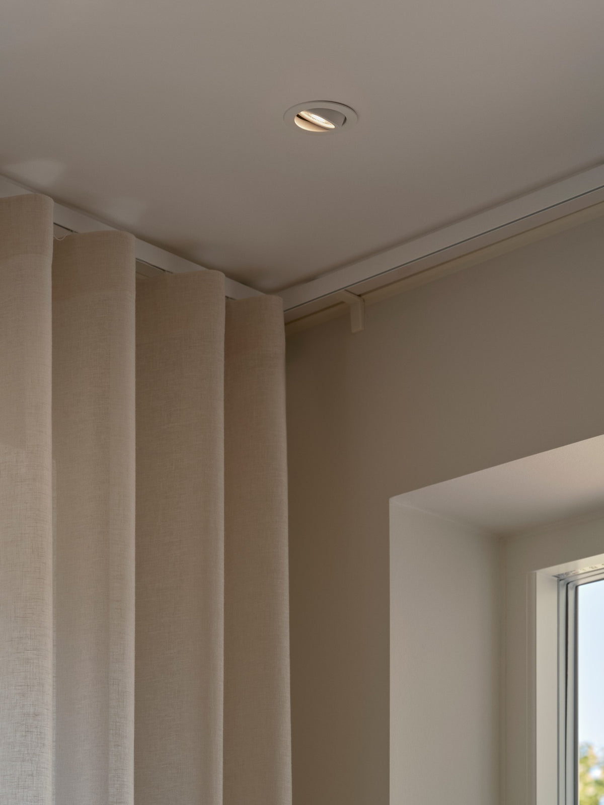 Mise en ambiance : spot encastré allumé au plafond près d'un rideau beige.
