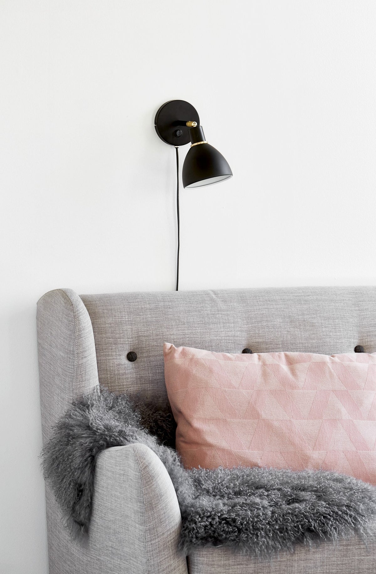 Applique murale noire fixée au-dessus d'un lit gris, coussin rose et plaid gris en ambiance chambre.
