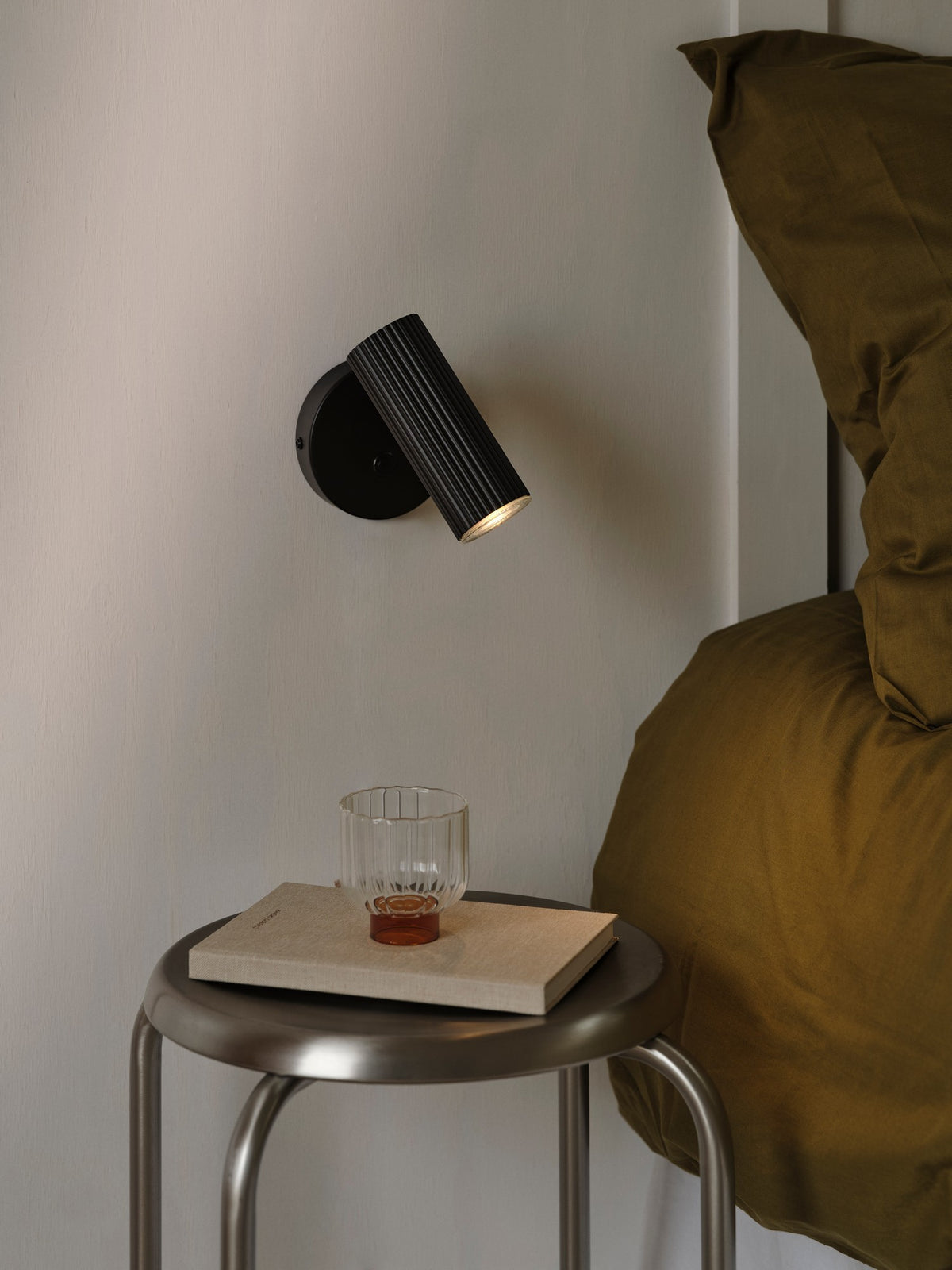 Mise en ambiance dans une chambre, applique murale noire allumée au-dessus d’une table de chevet.