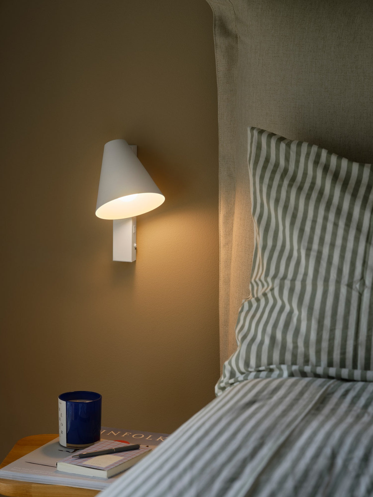 Mise en ambiance d'une chambre avec applique murale blanche allumée au-dessus d'une table de chevet.