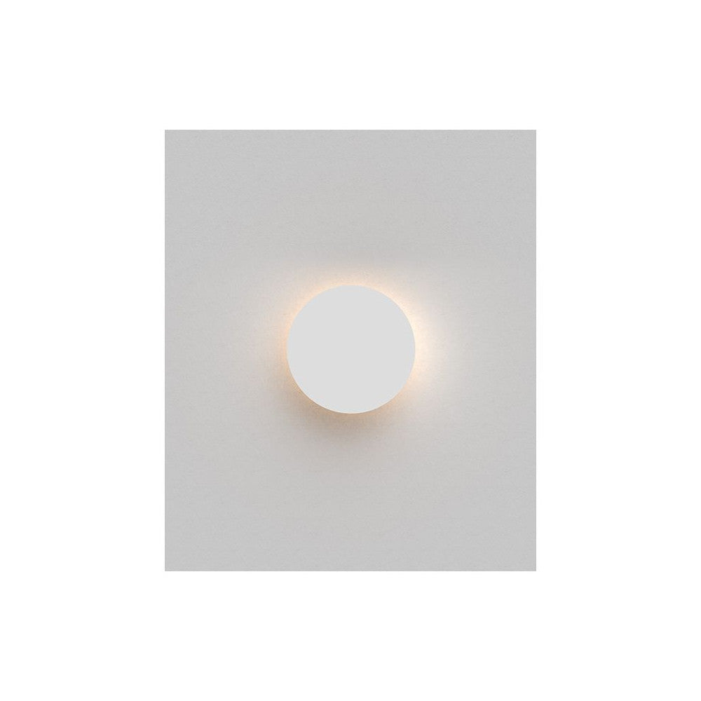 Applique murale ronde blanche allumée projetant un halo lumineux chaud sur mur gris