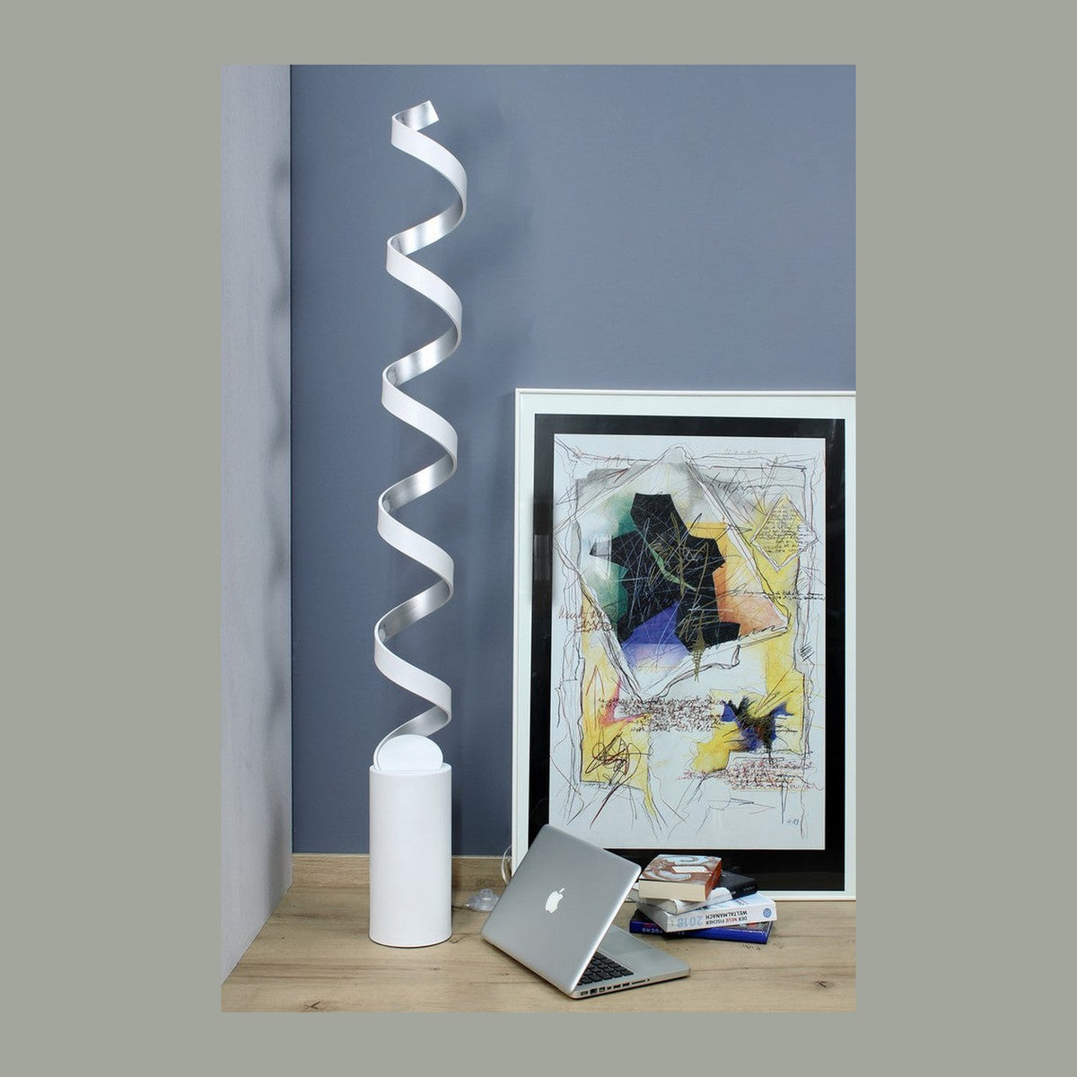 Mise en ambiance : lampadaire spiralé blanc près d'un mur gris, cadre, ordinateur et livres.