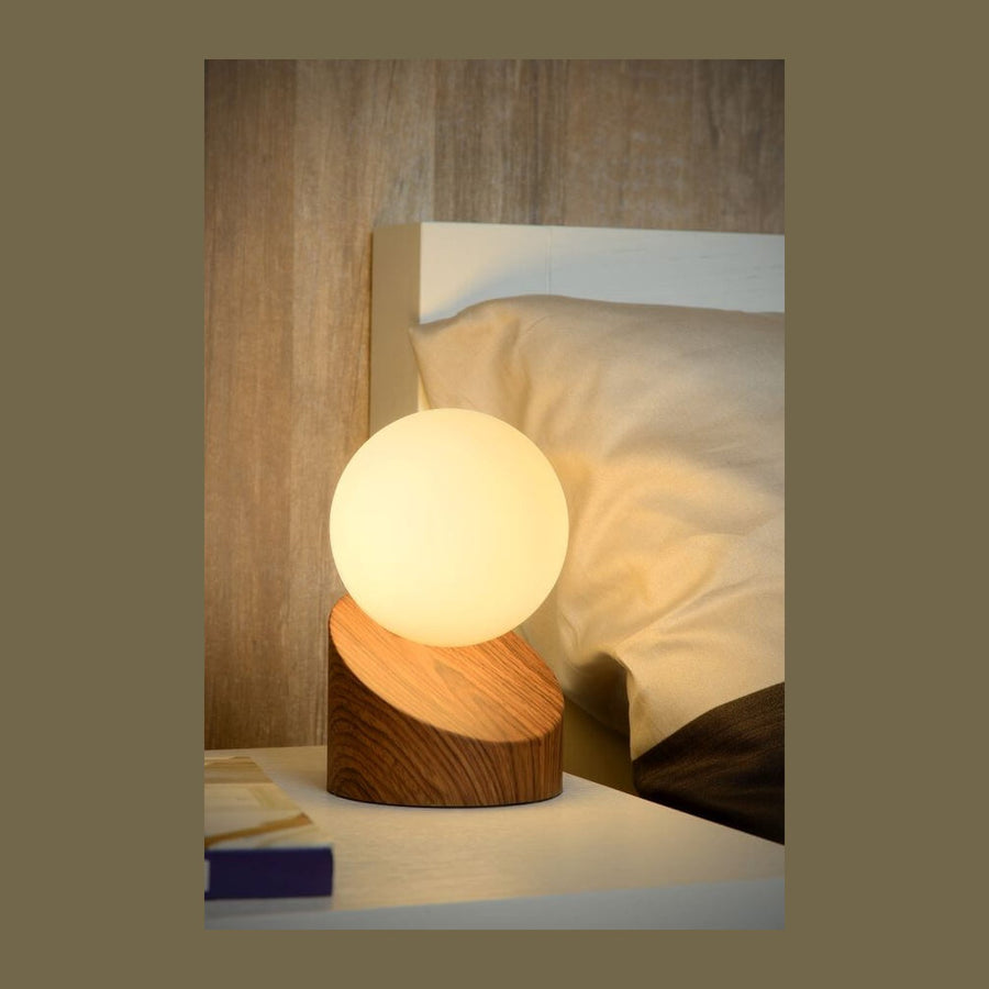 Lampe à poser en bois et sphère opaline allumée sur une table de chevet
