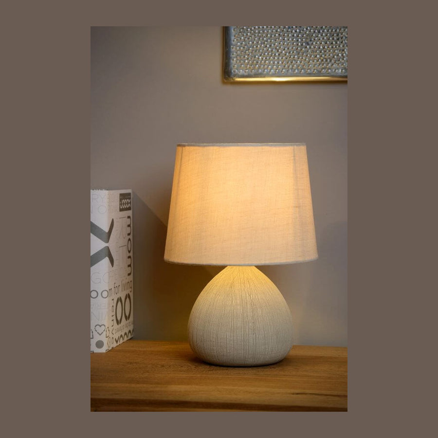 Lampe à poser beige allumée sur une commode en bois, avec boîte décorative à gauche.