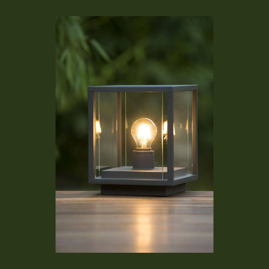 Luminaire cubique en verre allumé posé sur une table en bois, feuillage flou en arrière-plan.