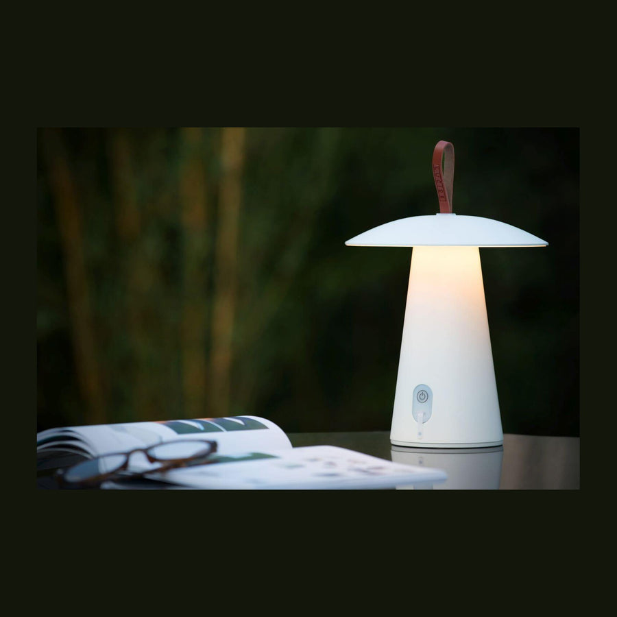 Lampe blanche allumée posée sur une table, avec magazine ouvert et lunettes devant un fond végétal.