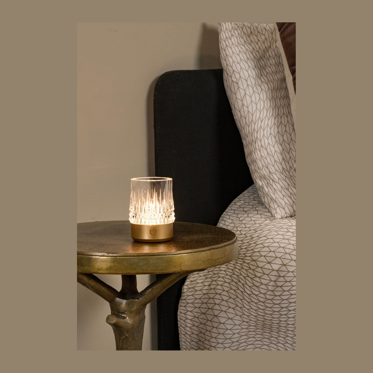 Lampe dorée allumée posée sur une table ronde devant un lit, ambiance chaleureuse.