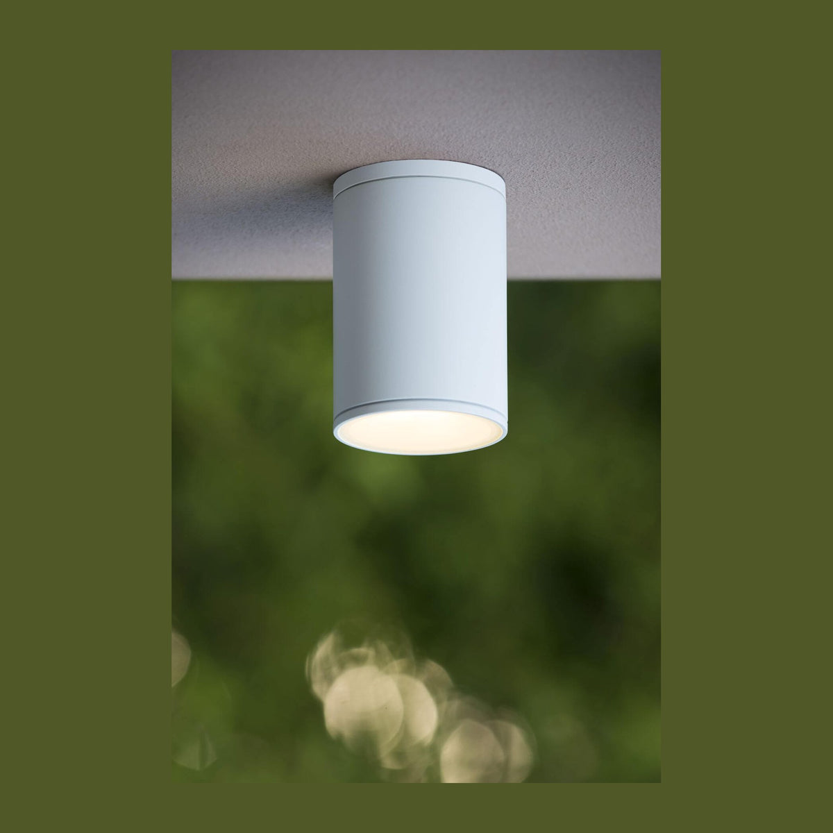 Plafonnier blanc cylindrique fixé au plafond extérieur, avec feuillage vert flou en arrière-plan.