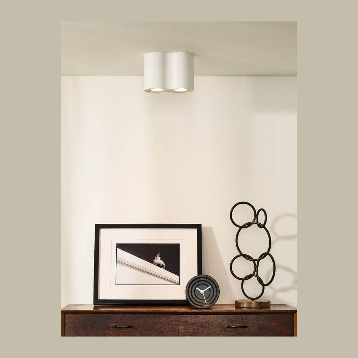 Plafonnier blanc bi-tube fixé au plafond, au-dessus d'un buffet en bois et d'une œuvre encadrée.