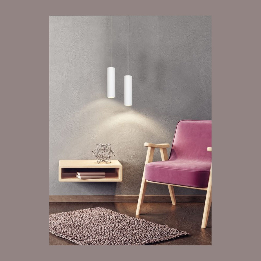 Suspension cylindrique blanche allumée éclairant un mur gris derrière un meuble et un fauteuil rose.