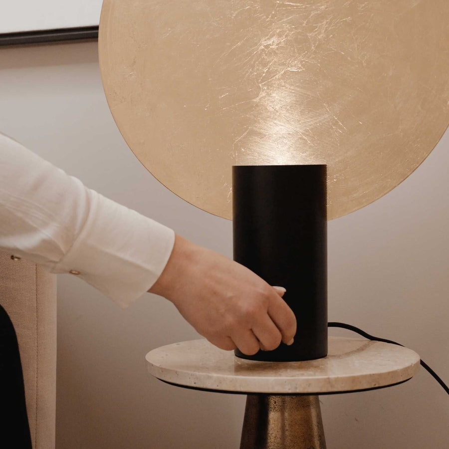 Main actionnant le socle de la lampe sur une table ronde en marbre.