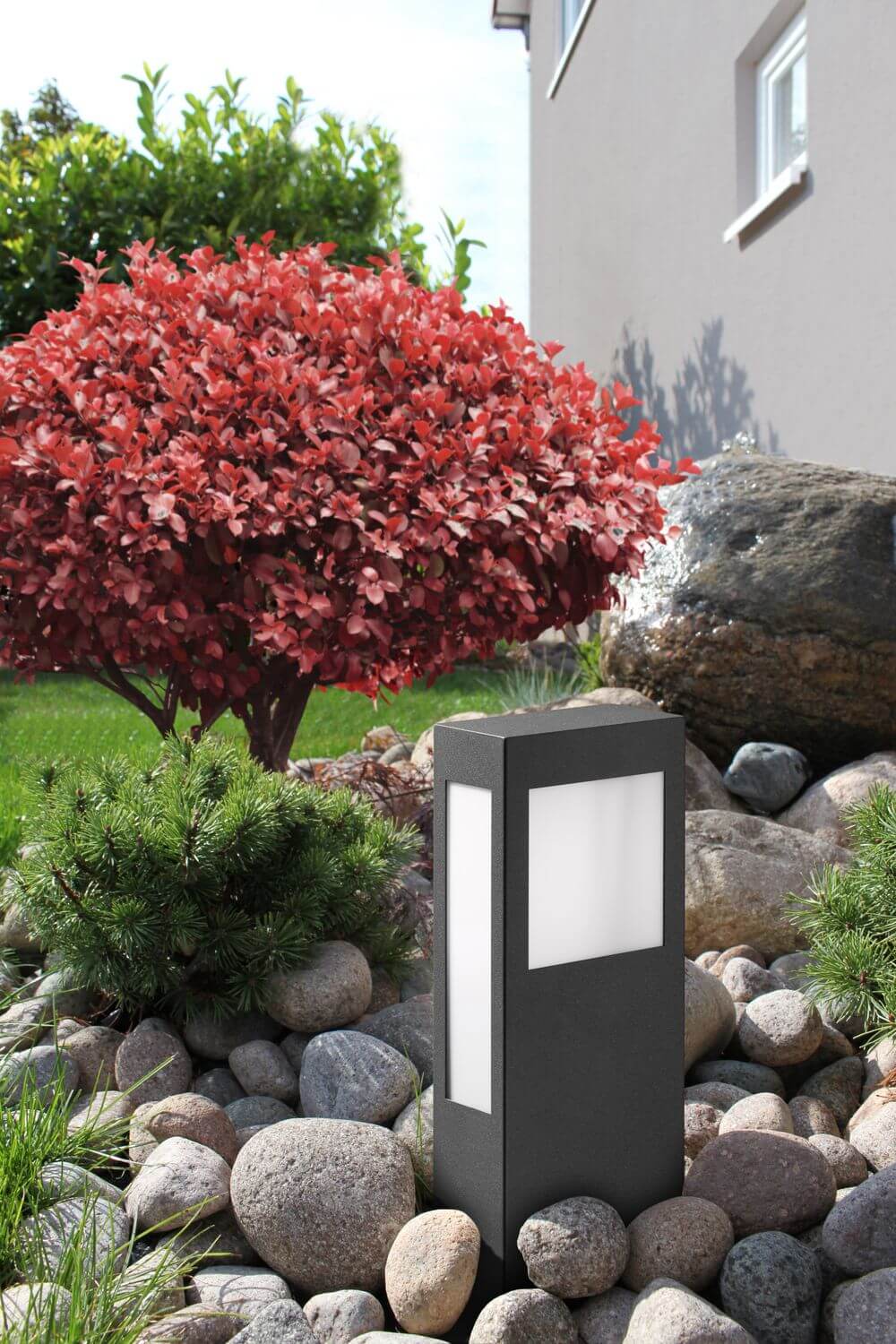 Mise en ambiance extérieure parmi des galets et arbustes, luminaire anthracite en situation.