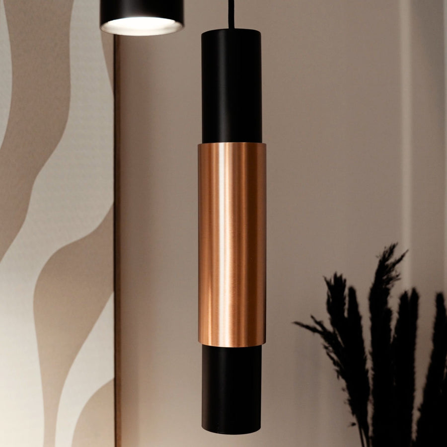 Gros plan sur une suspension cylindrique noire et cuivrée éclairant un mur beige.