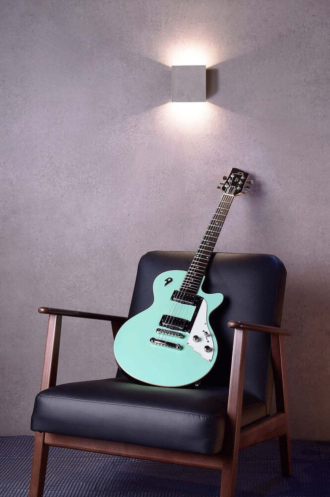 Mise en ambiance : applique murale allumée au-dessus d’un fauteuil noir et d’une guitare menthe.