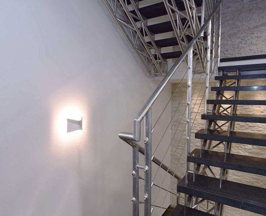 Applique murale allumée sur un mur d'escalier, halo lumineux et rampe métallique.