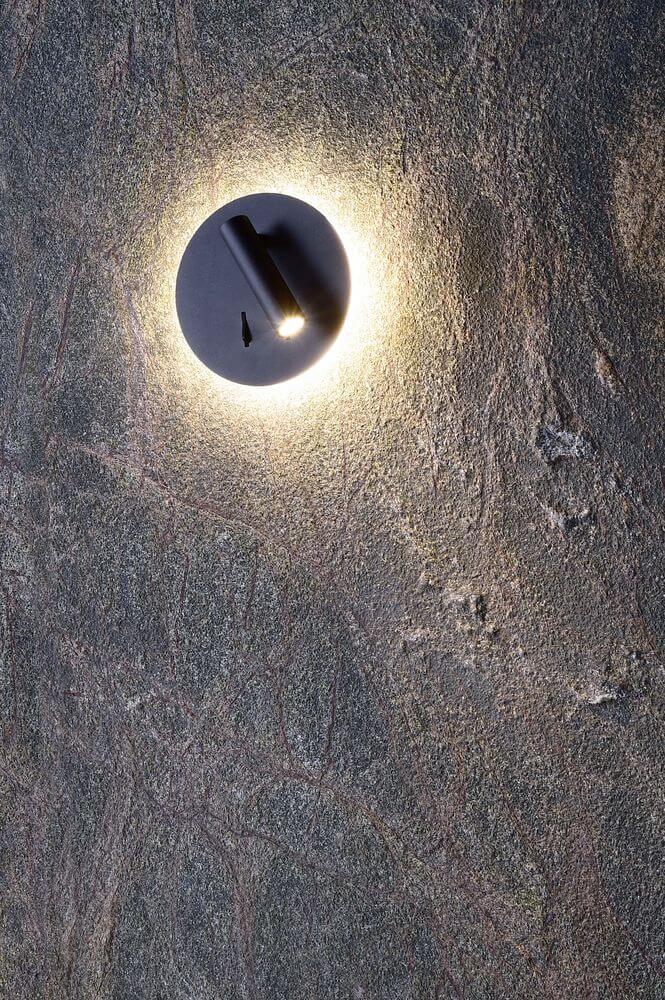 Applique murale allumée sur un mur texturé, halo lumineux diffus autour du socle rond.
