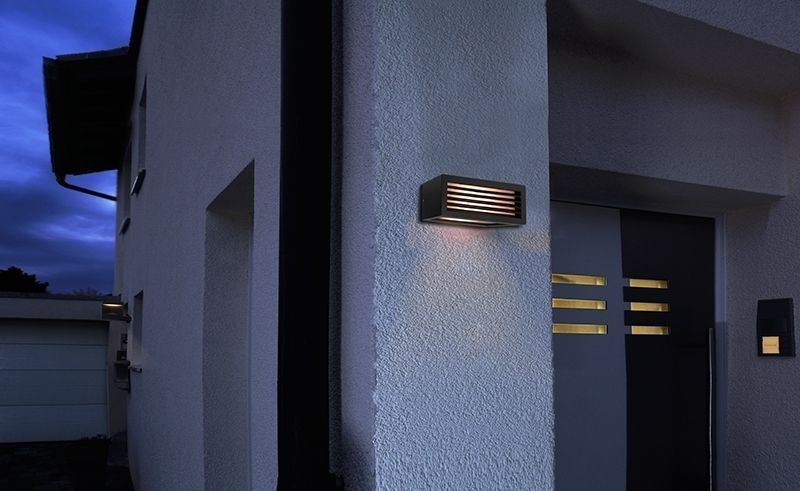 Mise en ambiance extérieure de l’applique allumée sur un mur blanc près d’une porte.