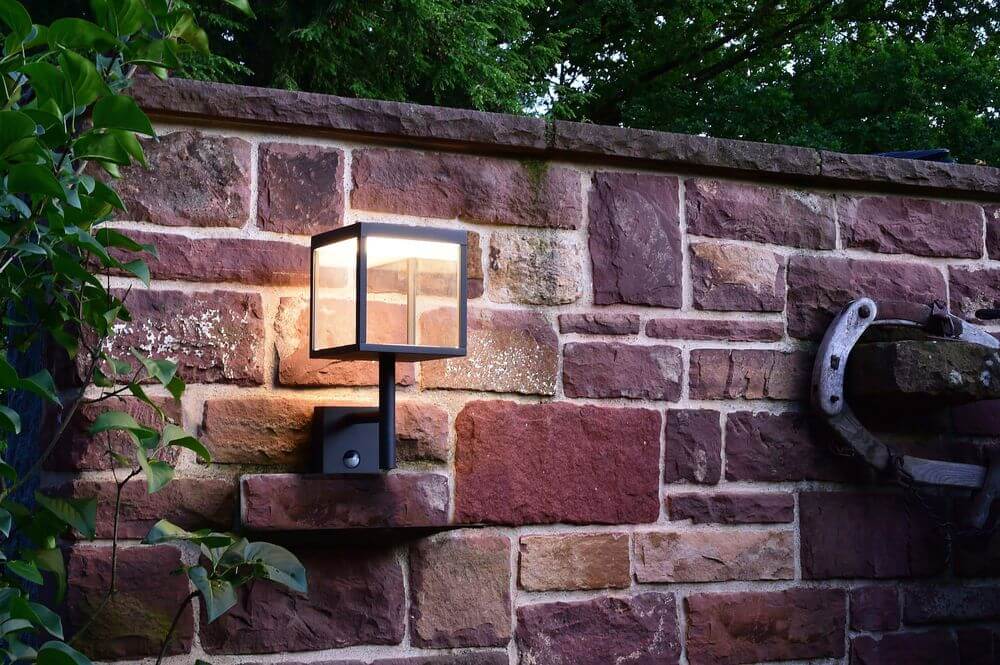Applique cube éclairée fixée sur un mur de pierres, ambiance de jardin au crépuscule.