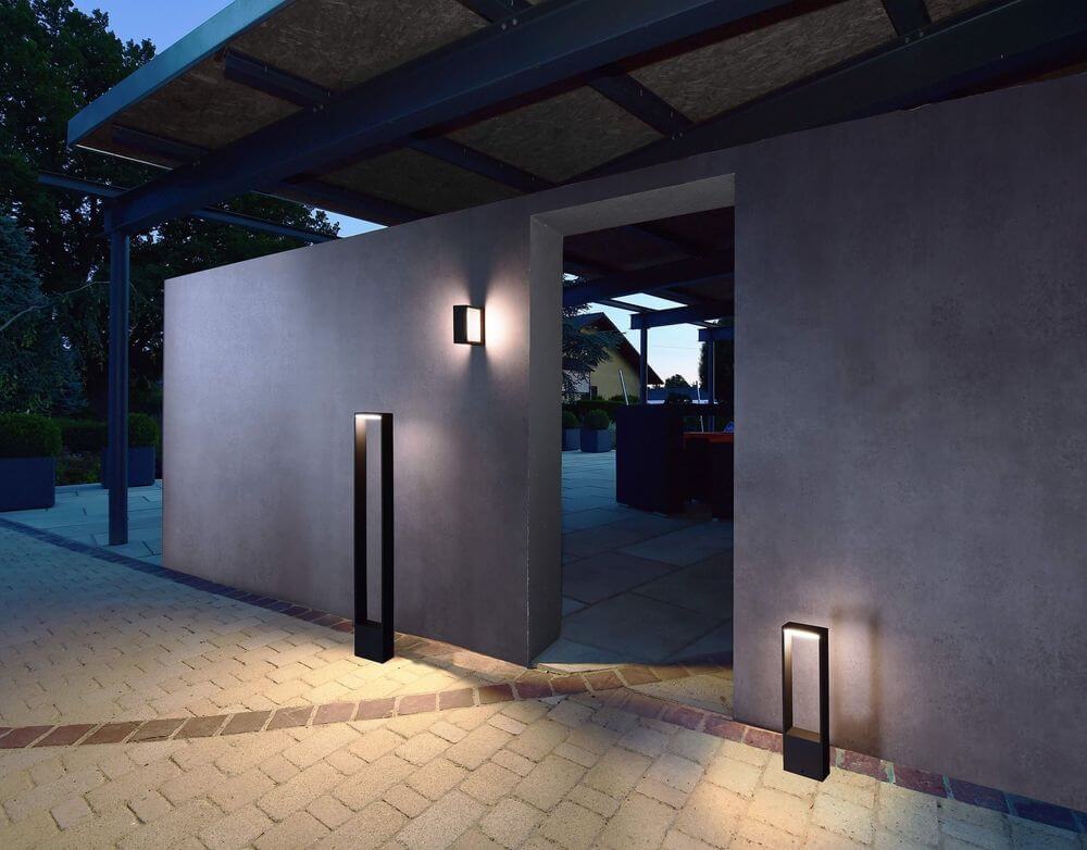 Mise en ambiance nocturne sur terrasse, lampadaire anthracite éclairant une allée pavée.