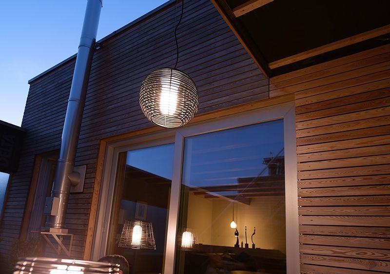 Suspension allumée installée devant une façade en bois, la cage sphérique diffusant une lumière chaude.