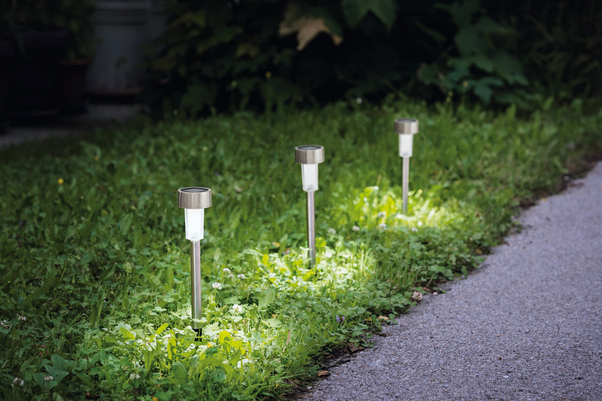 Trois luminaires solaires plantés dans l'herbe, éclairant une bordure près d'un chemin.