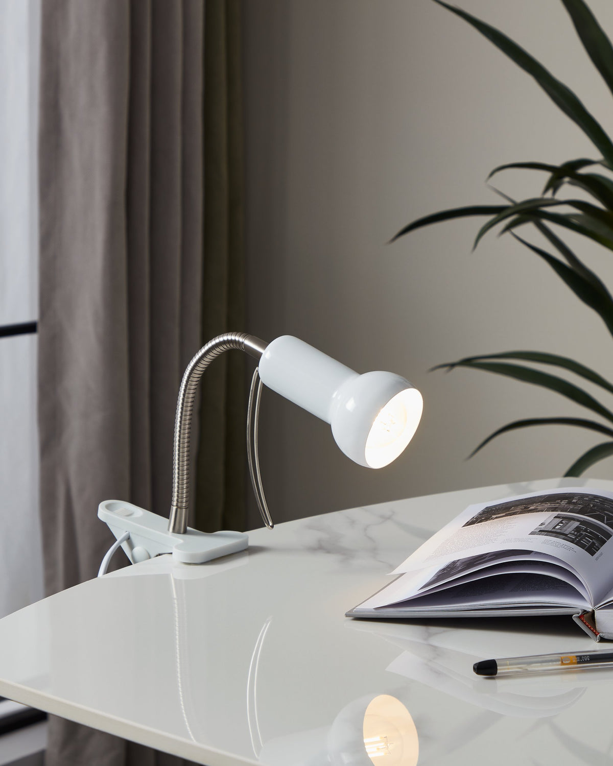 Lampe de bureau blanche allumée éclairant une revue ouverte sur un bureau avec plante.
