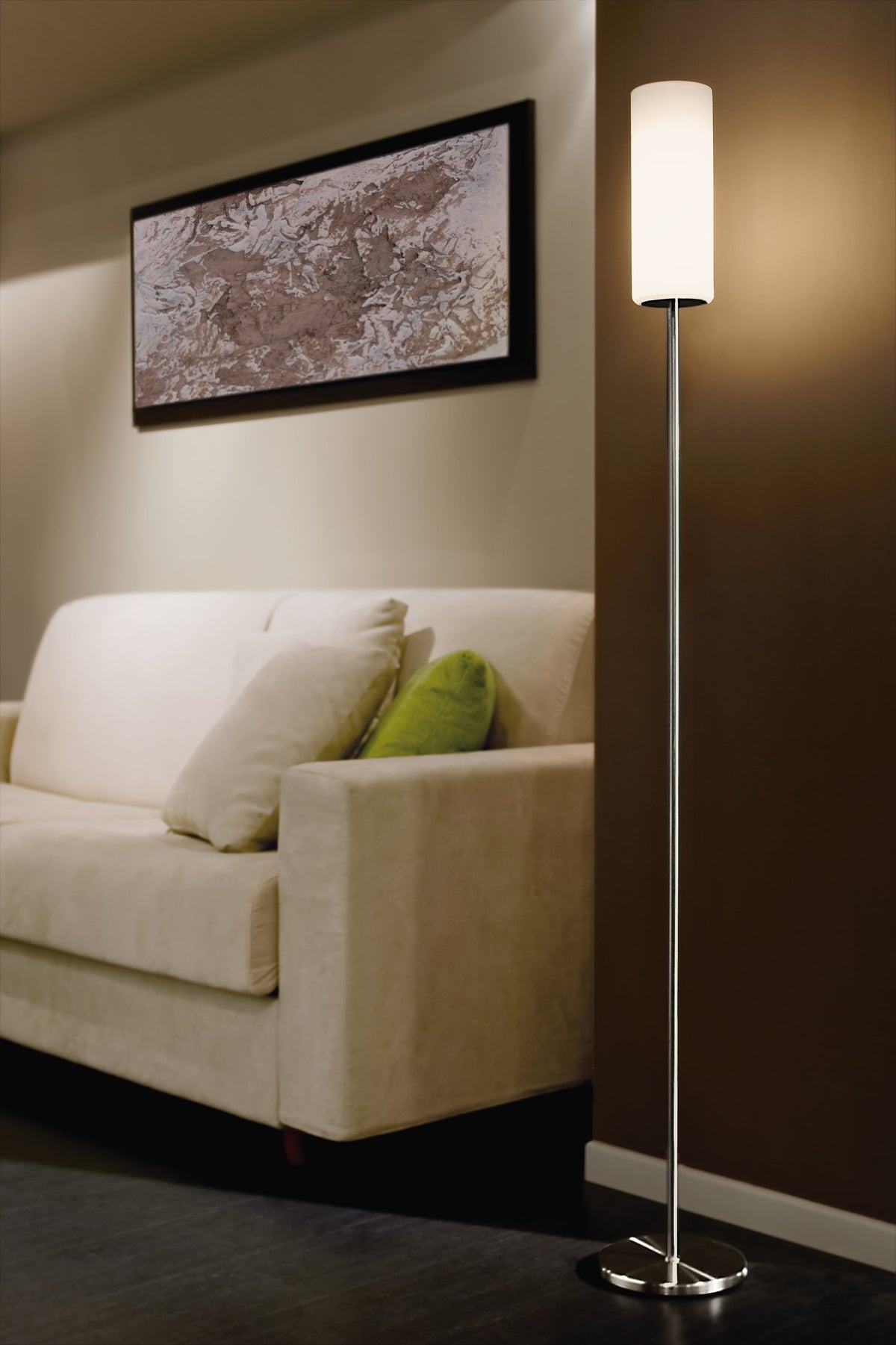 Mise en ambiance salon avec lampadaire allumé devant un canapé beige et un cadre.