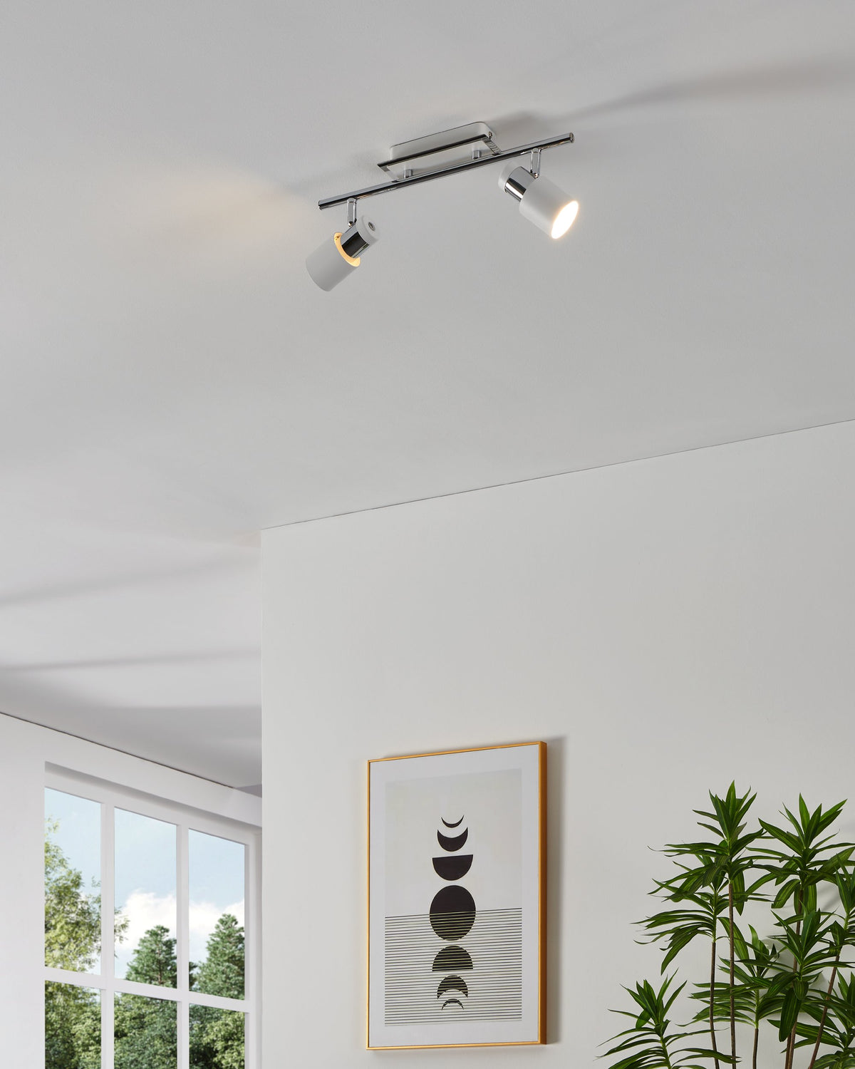 Luminaire spot double chromé et blanc fixé au plafond, éclairant un mur avec cadre et plante.