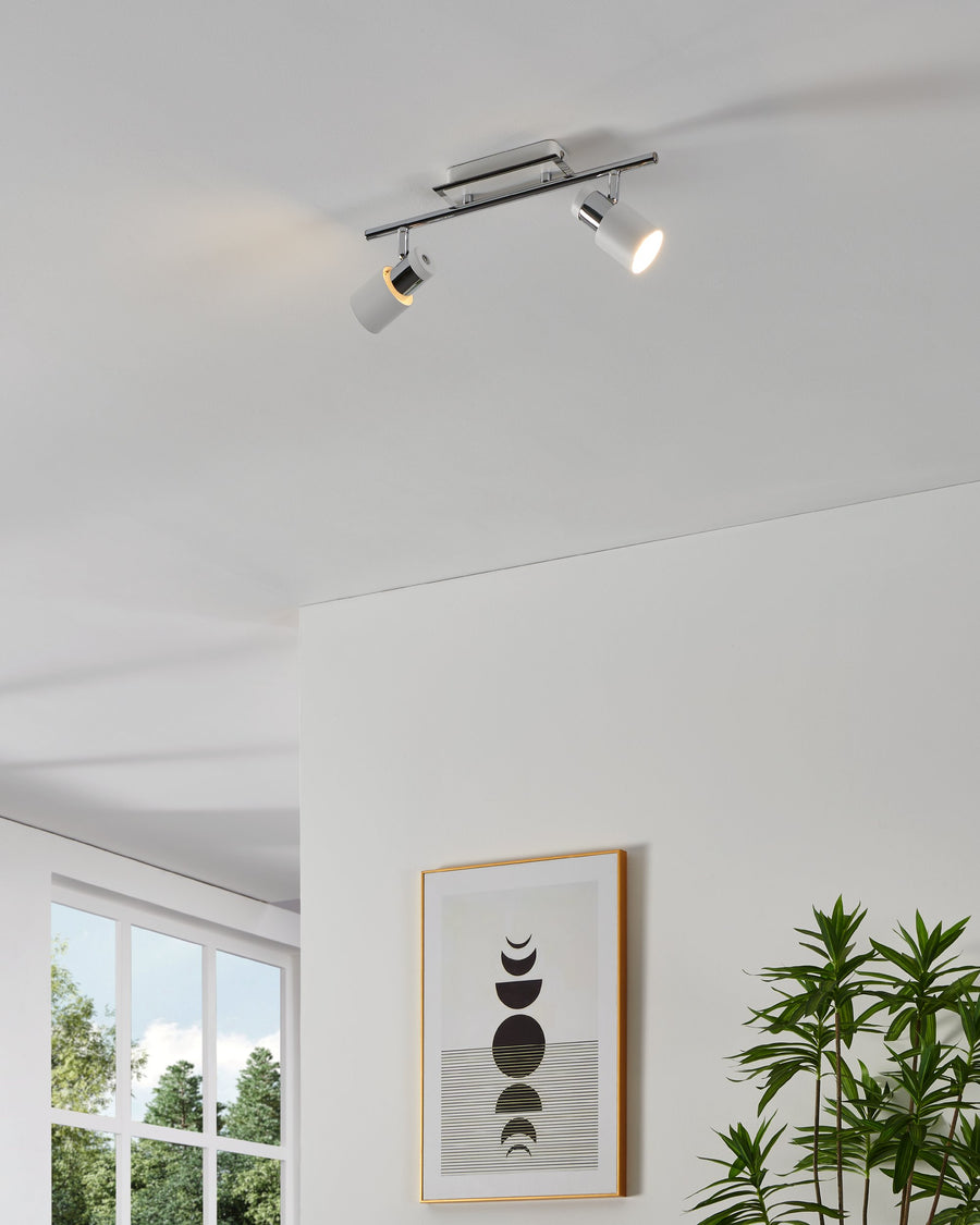 Luminaire spot double chromé et blanc fixé au plafond, éclairant un mur avec cadre et plante.