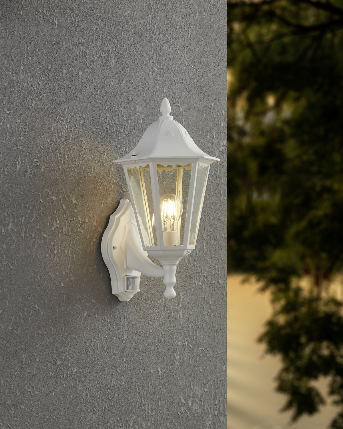Lanterne murale blanche allumée fixée sur un mur gris, feuillage flou en arrière-plan