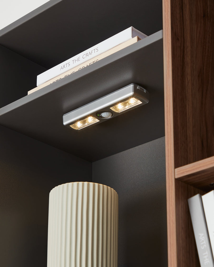Luminaire fixé sous une étagère sombre, éclairant livres et vase cannelé.