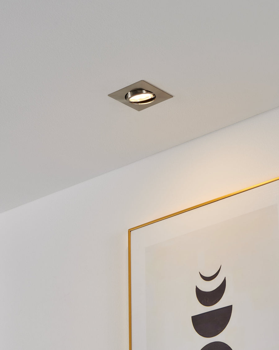 Spot encastré carré en nickel installé au plafond, éclairant un mur blanc et un cadre doré.